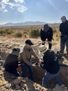 Three men standing in a trench working together to connect a water pipe, while 2 additional men overlook